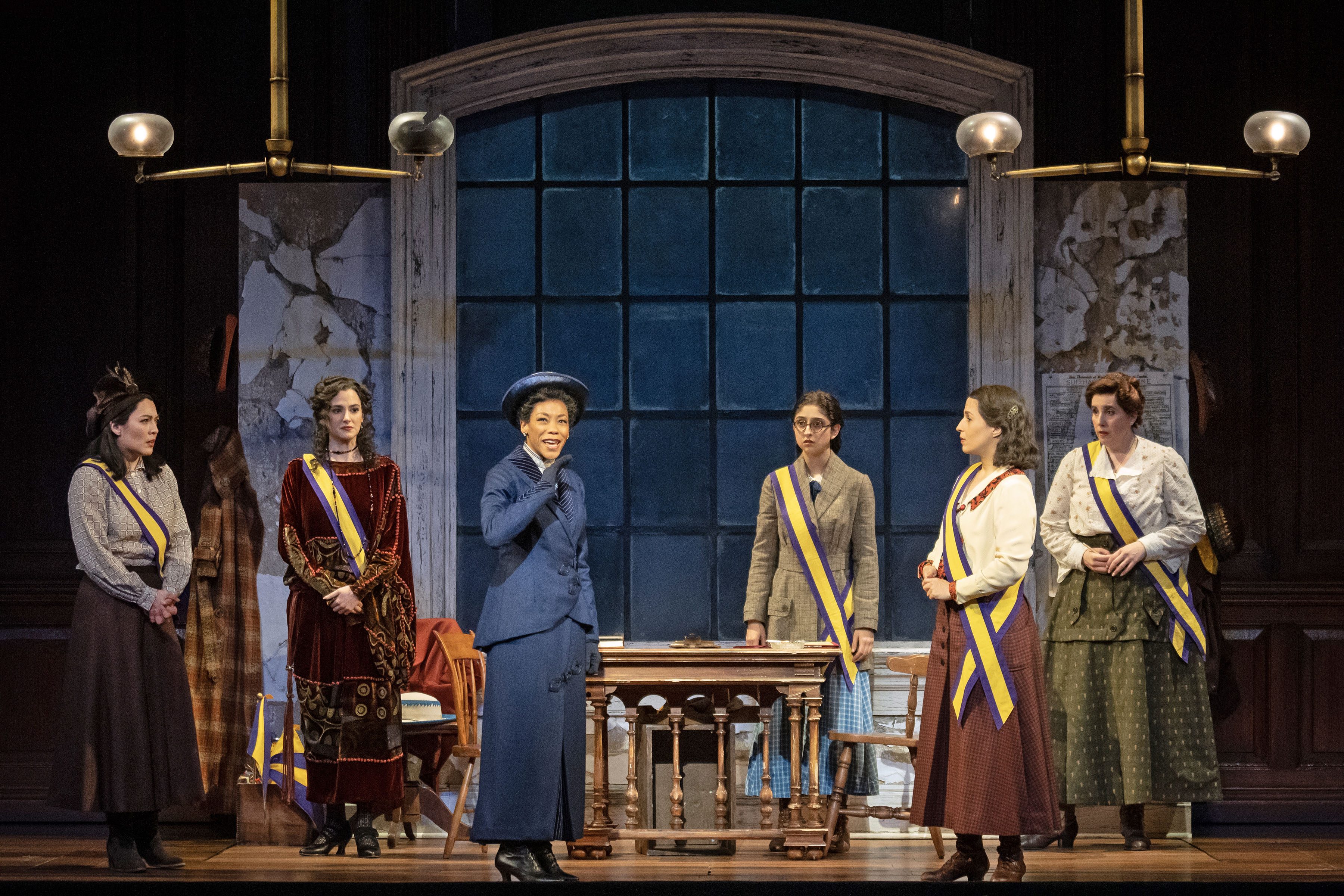 Six women in period costumes stand on stage with sashes in a historic setting. A woman in blue speaks confidently, suggesting a theme of empowerment.