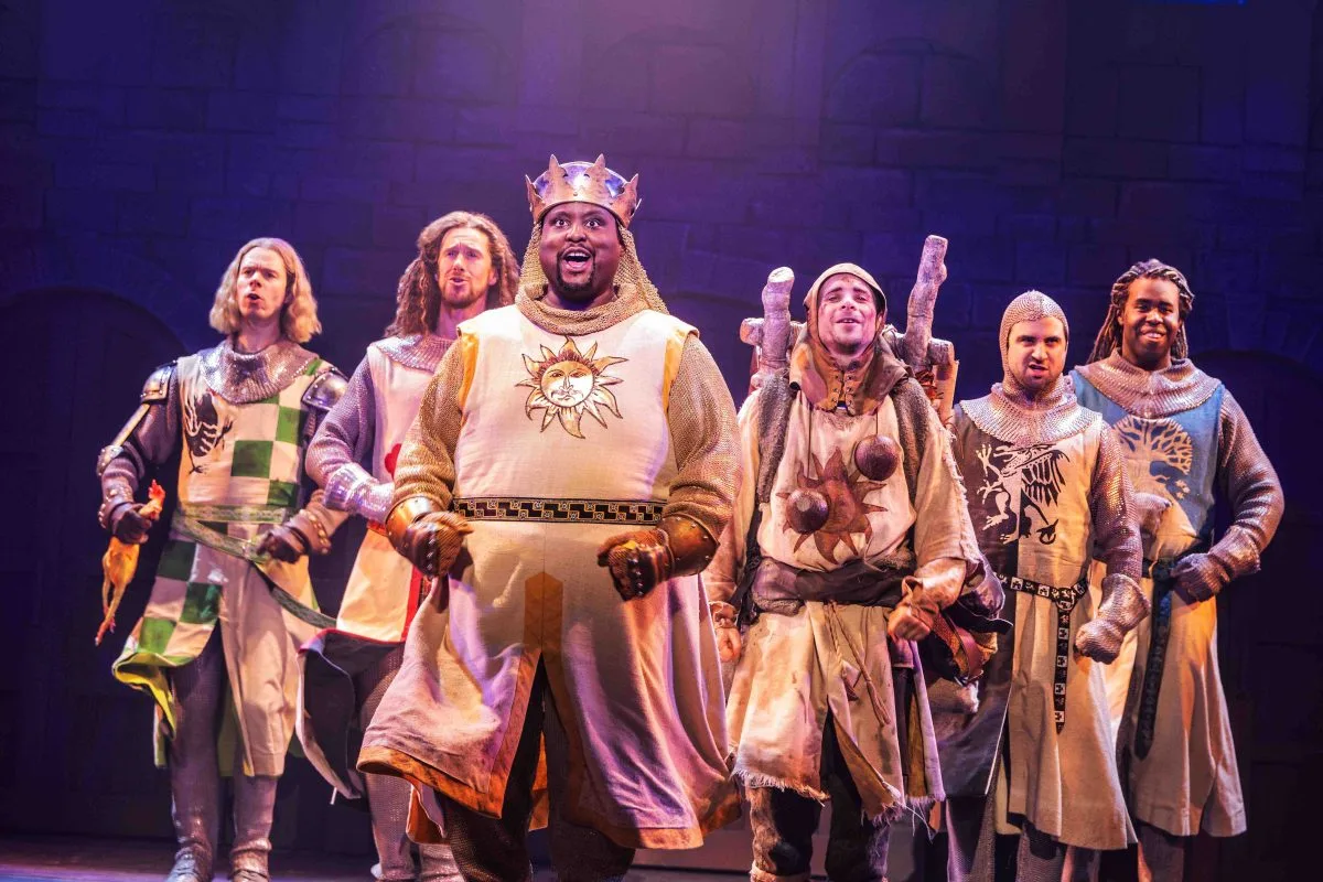 The cast of Spamalot dressed in knight costumes stand determined beneath a spotlight on their quest for the holy grail.